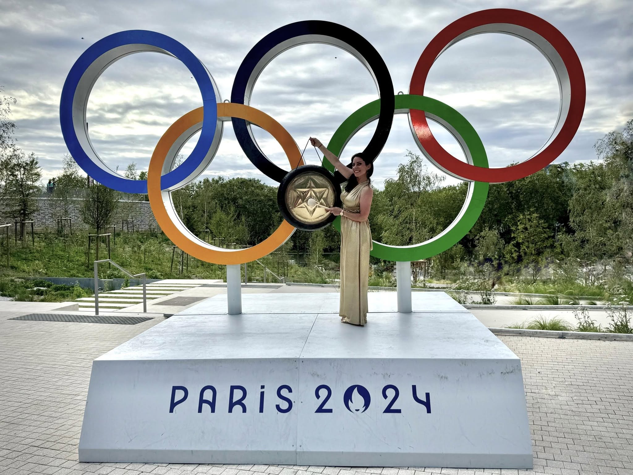Mélanie Zadeh devant les anneaux des Jeux Olympiques de Paris 2024 avec un gong